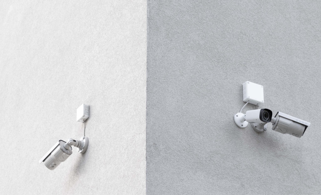 The exterior corner of a gray building with three white surveillance cameras.