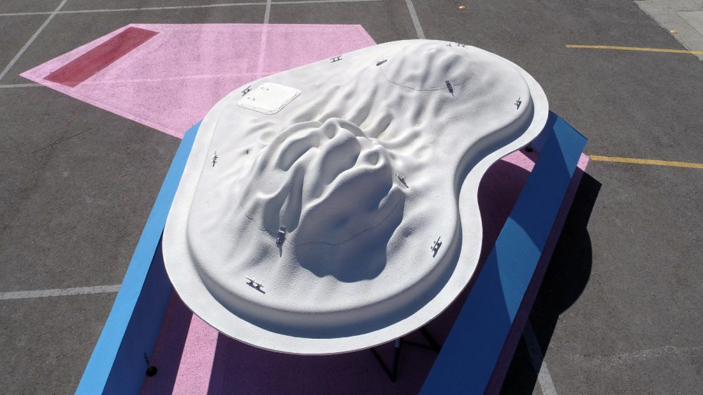 A white hull-like structure mounted in a blue display area on a parking lot painted with pink shapes.