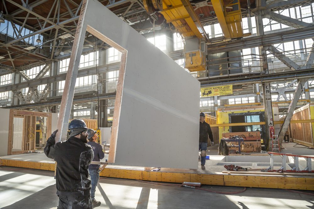 Workers carry a large segment of wall in a prefab housing factoring with a doorway cut out on a factory floor.