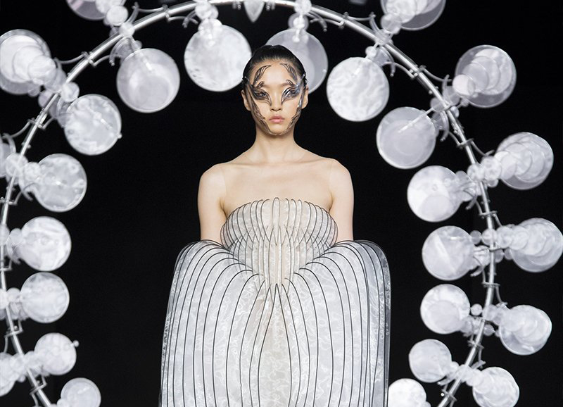 A model in a structured, widening dress comprising slits of fabric walks through a ring of rotating silk leaves designed by Iris van Herpen