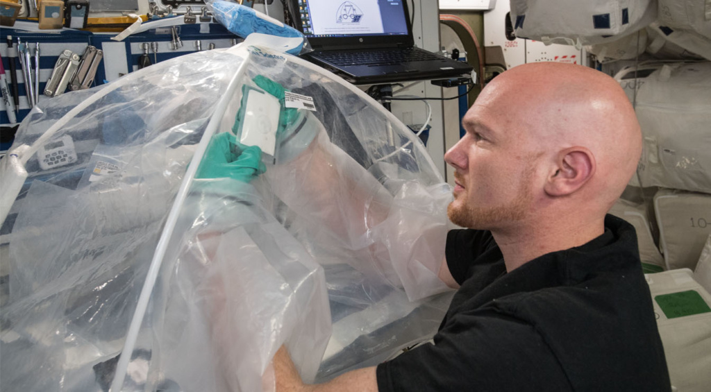 A person on a space station manipulating material inside a plastic apparatus to make concrete.