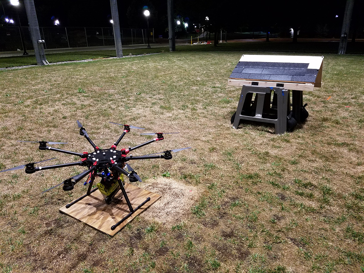 A drone on a lawn in front of a miniature roof on the ground.