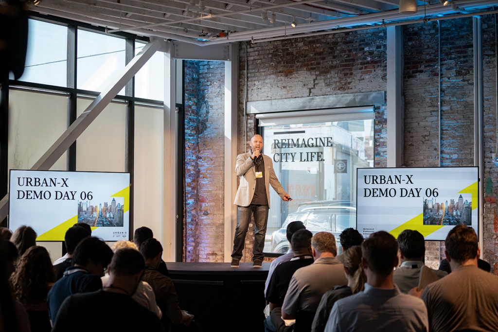 A person stands on a stage in front of two monitors while an audience looks on at Urban-X